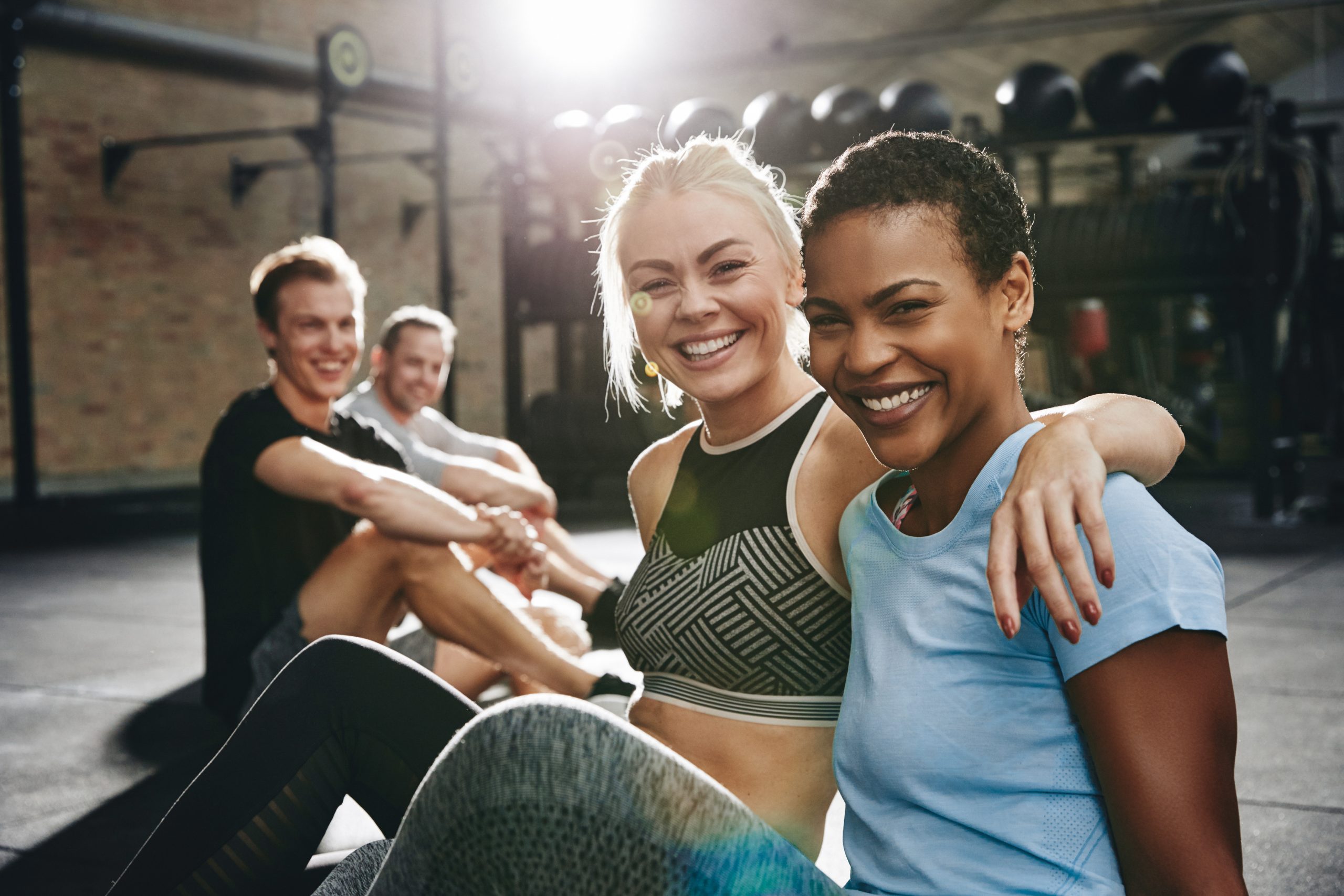 Two fit young women in sportswear laughing while sitting arm in arm together on the floor of a gym after working out